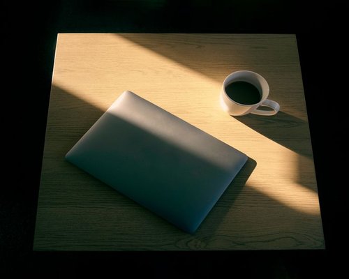 A laptop on a wooden desk near a window with soft morning sunlight casting linear shadows